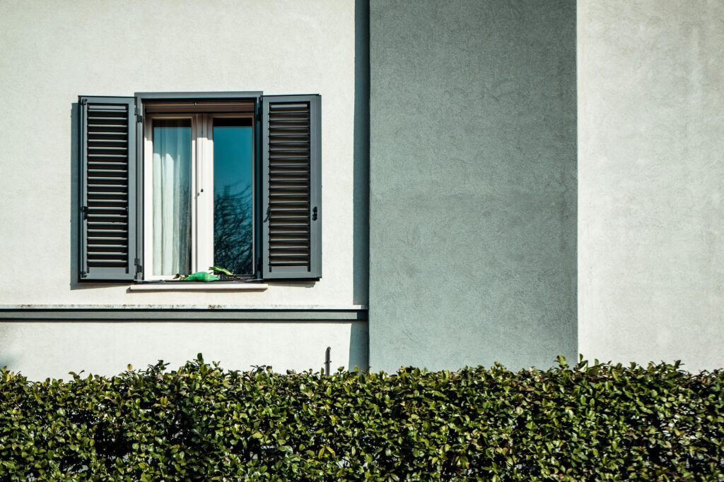 A minimalist exterior featuring a window with open shutters against a white wall with green hedges.